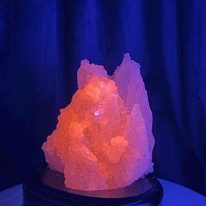 May include: A cluster of crystalline mineral formations emitting a bright, fluorescent orange glow under ultraviolet light. The jagged, translucent structure features numerous small, pointed points and sits atop a smooth, black, oval-shaped base.