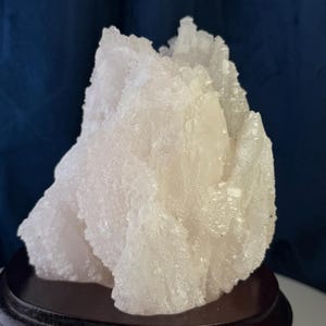 May include: A translucent, pale white mineral specimen with a jagged, crystalline surface texture. The cluster features numerous small, sharp points and irregular facets throughout its structure, mounted securely on a dark, circular wooden base.