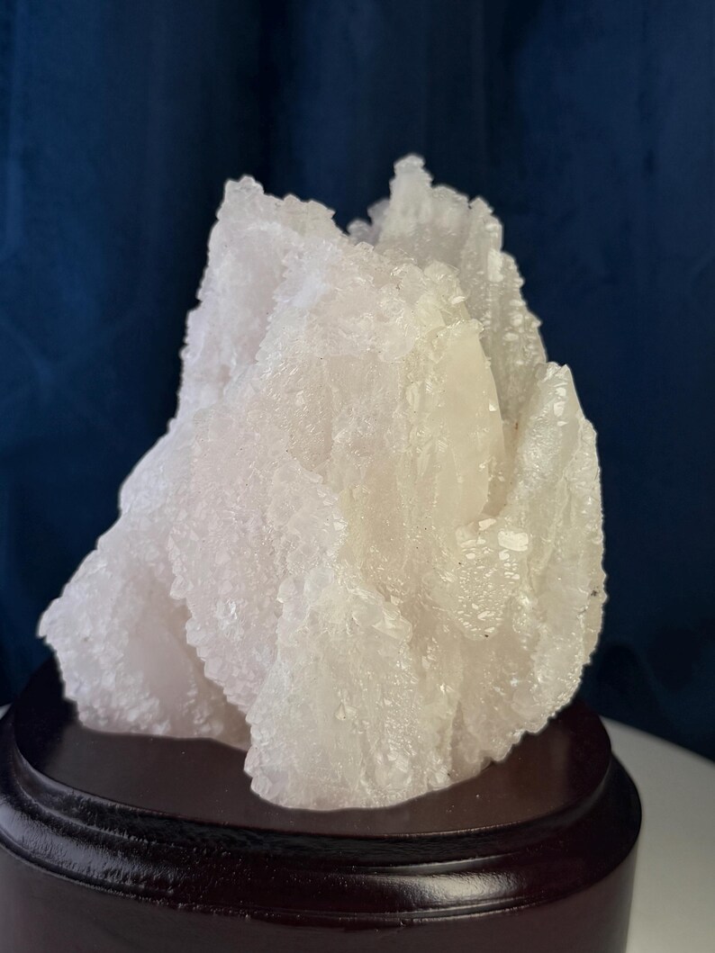 May include: A translucent, pale white mineral specimen with a jagged, crystalline surface texture. The cluster features numerous small, sharp points and irregular facets throughout its structure, mounted securely on a dark, circular wooden base.