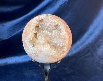 May include: A geode sphere with a hollow center filled with clear quartz crystals. The sphere is a light brown color and is sitting on a black metal stand.