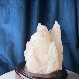 May include: A translucent, pale white crystalline mineral cluster with jagged, pointed formations. The mineral specimen is mounted on a circular, dark brown polished wooden base, showcasing a rough, textured surface with natural mineral facets.