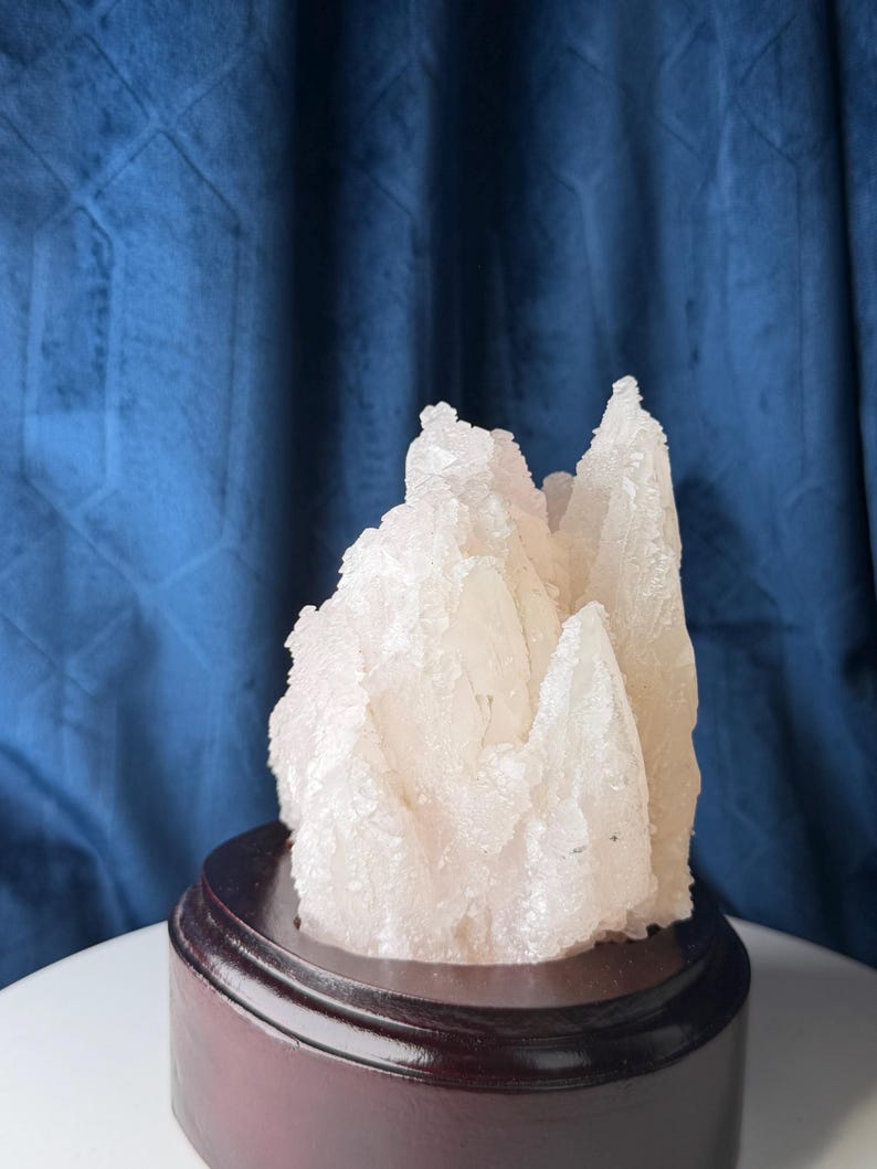 May include: A translucent, pale white crystalline mineral cluster with jagged, pointed formations. The mineral specimen is mounted on a circular, dark brown polished wooden base, showcasing a rough, textured surface with natural mineral facets.