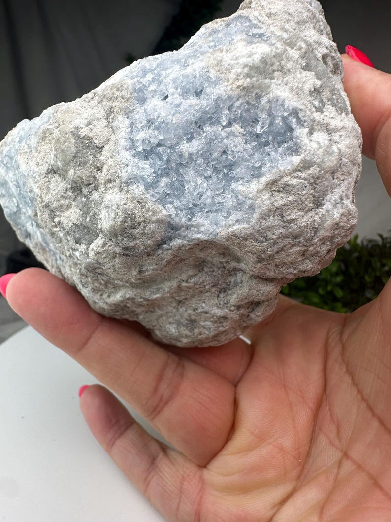 May include: A large celestite crystal, featuring a pale blue interior and a grey exterior. The rough-textured stone is held in a hand, highlighting its natural form and colour variations. The crystal appears to be a raw, uncut specimen.