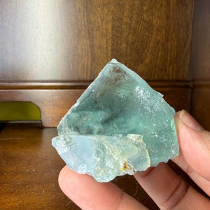 May include: A translucent, blue-green fluorite crystal with a rough, irregular shape. The crystal has a layered appearance and varying shades of color, with some areas appearing more clear than others. The edges are jagged, and the overall texture is slightly rough.