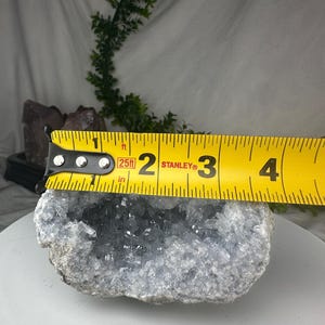 May include: A light blue celestite geode with a crystal-filled center. The geode is measured with a yellow tape measure, showing a width of approximately 7.6 cm. The geode has a rough, textured exterior.
