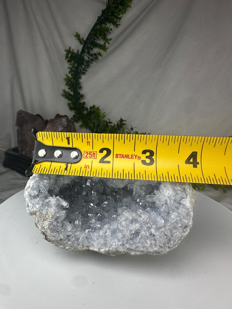 May include: A light blue celestite geode with a crystal-filled center. The geode is measured with a yellow tape measure, showing a width of approximately 7.6 cm. The geode has a rough, textured exterior.