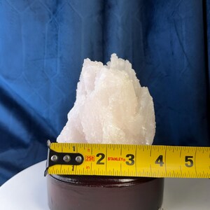 May include: A pale pink, crystalline mineral specimen with a jagged, pointed structure, mounted on a dark brown circular wooden base. A yellow Stanley measuring tape is positioned across the front, showing a width of approximately 8.9 centimetres.