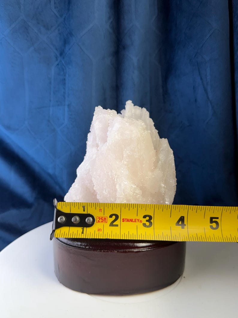 May include: A pale pink, crystalline mineral specimen with a jagged, pointed structure, mounted on a dark brown circular wooden base. A yellow Stanley measuring tape is positioned across the front, showing a width of approximately 8.9 centimetres.