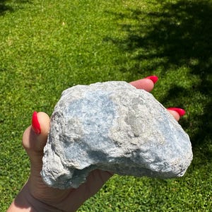 May include: A pale blue, textured stone held in a hand with red nail polish. The stone has a mix of light blue and grey tones. The background is a green lawn.