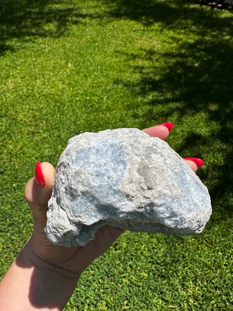 May include: A pale blue, textured stone held in a hand with red nail polish. The stone has a mix of light blue and grey tones. The background is a green lawn.