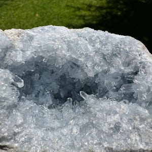 May include: A large, pale blue celestite geode with a crystalline interior. The geode's outer layer is a rough, grey-white colour, contrasting with the sparkling, translucent crystals inside. The crystals are clustered, creating a textured, natural display.