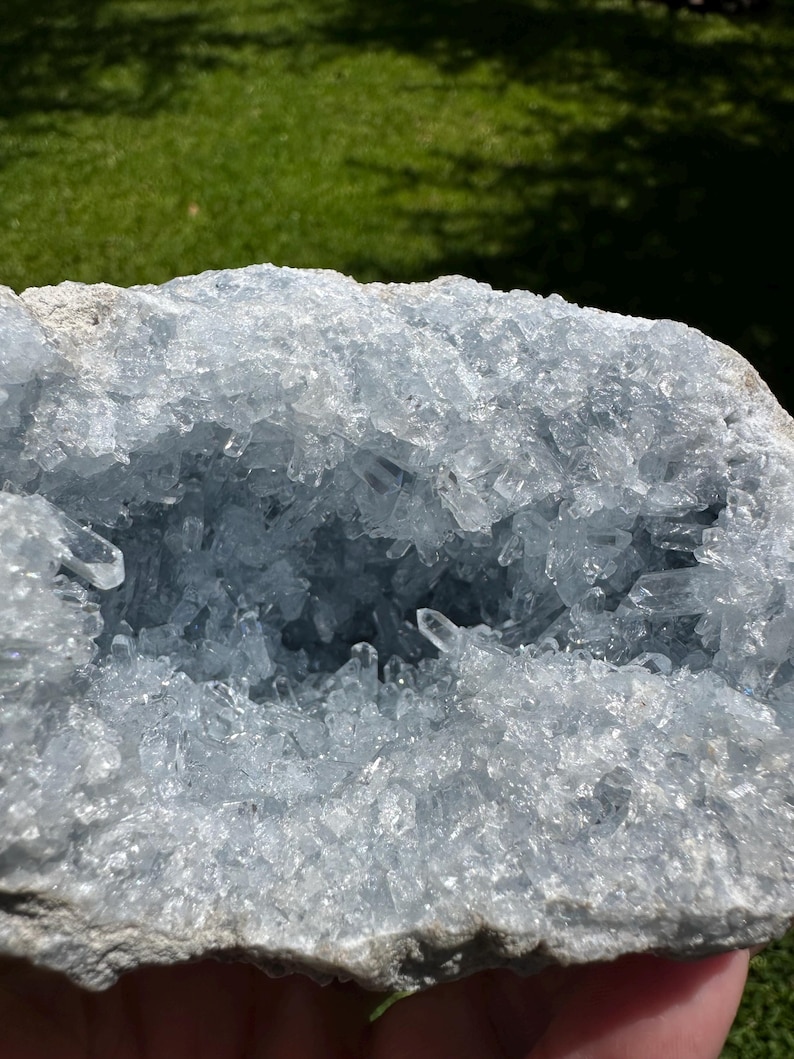 May include: A large, pale blue celestite geode with a crystalline interior. The geode's outer layer is a rough, grey-white colour, contrasting with the sparkling, translucent crystals inside. The crystals are clustered, creating a textured, natural display.