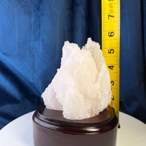May include: A white, crystalline mineral specimen with a jagged, textured surface mounted on a dark brown, circular wooden base. A yellow measuring tape is positioned vertically next to the crystal, showing a height of approximately 8.9 centimetres.