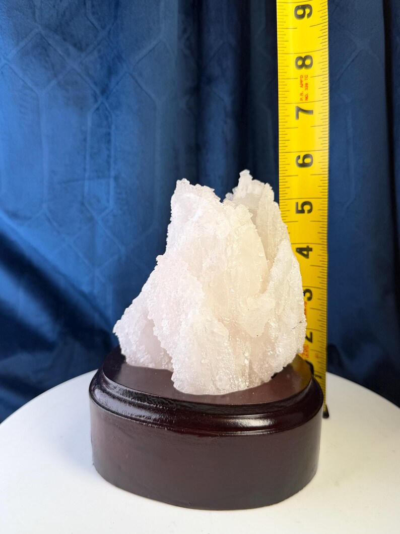 May include: A white, crystalline mineral specimen with a jagged, textured surface mounted on a dark brown, circular wooden base. A yellow measuring tape is positioned vertically next to the crystal, showing a height of approximately 8.9 centimetres.