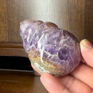 May include: A polished amethyst crystal sculpture in shades of purple, brown, and white. The crystal is held in a hand and has a rounded, organic shape. The sculpture is displayed against a dark wooden background.