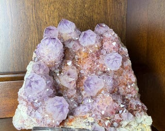 May include: A cluster of amethyst crystals in shades of purple and pink, mounted on a clear acrylic stand. The crystals have a sparkling, natural appearance, with a rough, textured base. The overall shape is irregular and organic.