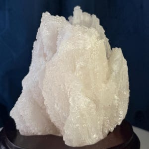 May include: A translucent white mineral specimen with a jagged, crystalline texture and irregular peaks. The cluster is mounted on a smooth, circular, dark brown wooden base, providing a stable foundation for the rough, naturally formed stone structure.