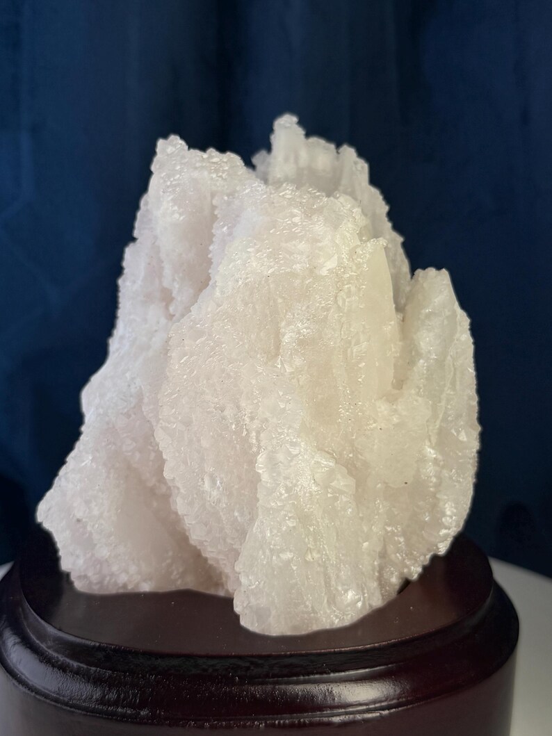 May include: A translucent white mineral specimen with a jagged, crystalline texture and irregular peaks. The cluster is mounted on a smooth, circular, dark brown wooden base, providing a stable foundation for the rough, naturally formed stone structure.