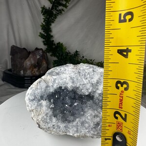 May include: A geode with a light blue crystal interior and a rough, grey exterior. The geode is approximately 7.6 cm in diameter, as measured by a yellow Stanley tape measure. The geode is a natural mineral specimen.
