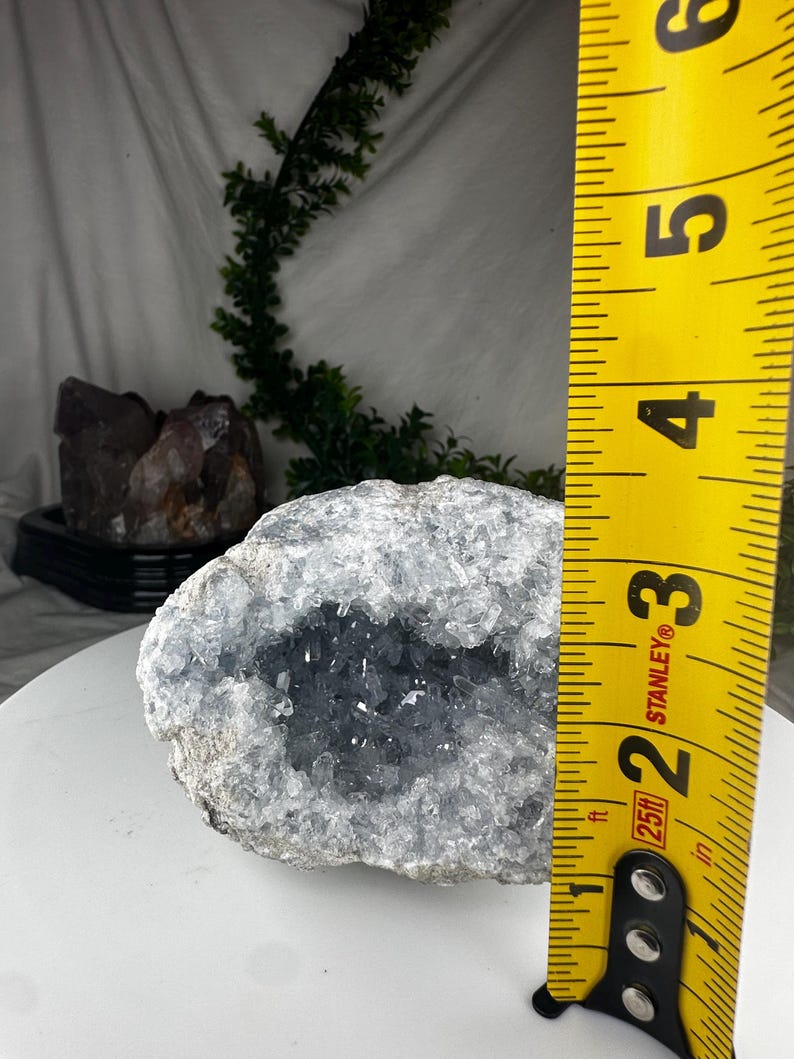 May include: A geode with a light blue crystal interior and a rough, grey exterior. The geode is approximately 7.6 cm in diameter, as measured by a yellow Stanley tape measure. The geode is a natural mineral specimen.