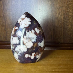 May include: A polished, teardrop-shaped stone with a dark purple base and white, flower-like patterns. The stone is smooth and glossy, with a natural, organic appearance. It sits on a wooden surface, showcasing its unique color and texture.