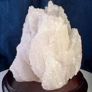 May include: A translucent, white crystalline mineral specimen with a jagged, irregular surface texture. The cluster features numerous small, reflective crystal points and is mounted on a smooth, circular, dark brown polished wooden base.