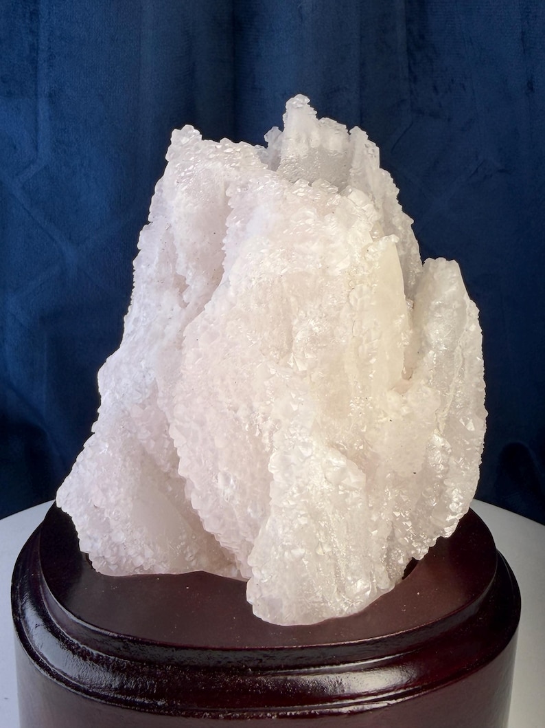 May include: A translucent, white crystalline mineral specimen with a jagged, irregular surface texture. The cluster features numerous small, reflective crystal points and is mounted on a smooth, circular, dark brown polished wooden base.