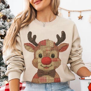 May include: Beige Christmas sweatshirt with a reindeer design, featuring a red nose, brown antlers, and a bow tie. The reindeer is made of patchwork fabric. A white mug with a Santa Claus illustration is held.