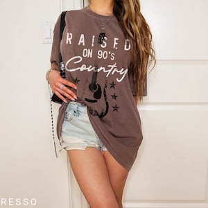 May include: An oversized brown t-shirt with a white graphic print of a guitar and the text "Raised on 90's Country".