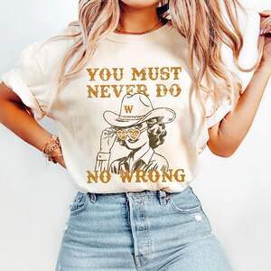 May include: A white t-shirt with the text "You must never do no wrong" and a drawing of a woman wearing a cowboy hat and sunglasses with the text "W" on the hat. The text is in brown and the drawing is in black and brown.