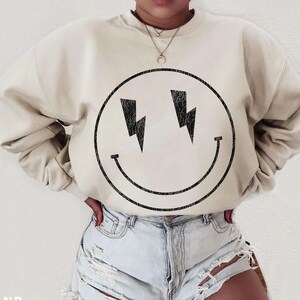 May include: A cream colored sweatshirt with a black smiley face graphic with lightning bolts for eyes. The model is wearing the sweatshirt with denim shorts.