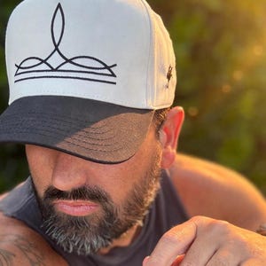 Könnte beinhalten: Eine Baseballkappe mit weißer Krone und schwarzem Schirm, die auf der Vorderseite eine schwarze Strichgrafik einer Krone aufweist. Die Kappe wird mit einem dunkelgrauen ärmellosen Shirt kombiniert, das weißen Text auf der Brust trägt.
