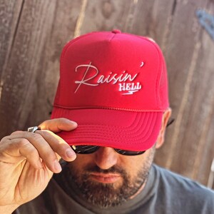 Puede incluir: Una gorra de camionero roja con un logotipo bordado en blanco que dice "Raisin' Hell". La persona que lleva la gorra también lleva una camiseta gris con el texto "Put Me In Coach" impreso en blanco.