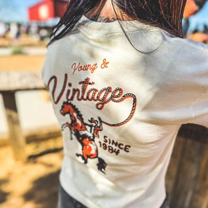 May include: Cream-colored long-sleeved shirt with the words "Young & Vintage" in orange script and a graphic of a rearing horse. The text "Since 1984" is below the horse graphic. The shirt is worn by a person.