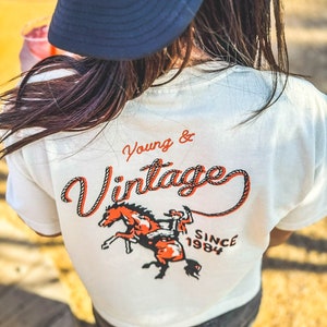May include: Cream-colored t-shirt with the words "Young & Vintage" in orange and a graphic of a cowboy riding a horse. The shirt also has the text "SINCE 1984". A black trucker hat with white text is worn.