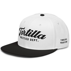 Puede incluir: Gorra de béisbol blanca con visera negra. La gorra presenta la palabra "Tortilla" en fuente cursiva, junto con "Analytics Dept." y "Timeless Tradition" impresos en negro.