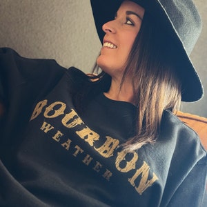 May include: A black sweatshirt with the text "Bourbon Weather" printed in gold on the front. The sweatshirt is worn by a person wearing a black hat.