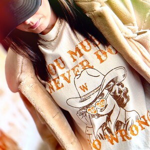 May include: A white t-shirt with the text "You must never do wrong" in orange lettering. The shirt features a cartoon illustration of a woman wearing a cowboy hat and sunglasses with the text "Pro Beer" on the lenses. The woman is holding a cigarette in her hand.