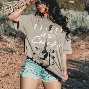 May include: A woman wearing a khaki t-shirt with the text "Raised on 90's Country" and a guitar graphic. She is wearing denim shorts and brown cowboy boots. The photo was taken outdoors in a desert setting.