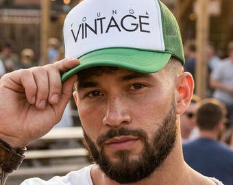 Può includere: Cappellino da camionista bianco e verde con la scritta "VINTAGE" stampata sul davanti. Il cappellino ha un retro in rete e una visiera curva. Il cappellino è tenuto da una persona.