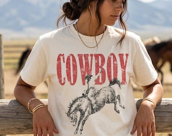 May include: A cream-coloured short-sleeve t-shirt featuring the red text COWBOY in a distressed font above a black and white graphic illustration of a person riding a bucking horse. The shirt has a crew neckline and a relaxed fit.