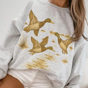 May include: A light gray sweatshirt featuring a graphic of three ducks in flight, with golden-brown feathers and yellow beaks. The ducks are set against a backdrop of golden grass. The sweatshirt is paired with white shorts.