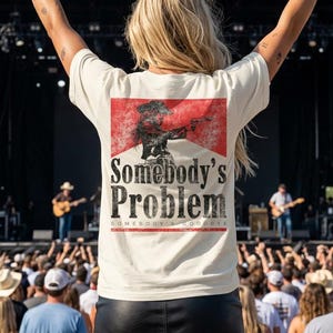 Può includere: Una t-shirt a maniche corte color crema con una grafica rettangolare rossa sul retro. La grafica raffigura la sagoma di una persona con un cappello da cowboy che impugna una pistola, con la scritta "Somebody's Problem" stampata in nero.