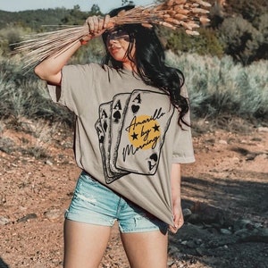 May include: A woman wearing a khaki t-shirt with a graphic of playing cards and the text "Amarillo by Morning". She is wearing denim shorts and cowboy boots. The photo was taken outdoors in a desert setting.