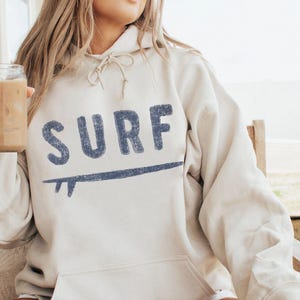 Puede incluir: Una sudadera con capucha de color arena con capucha con cordón y bolsillo de canguro. La palabra "SURF" está impresa en letras azules en el pecho, con un gráfico de tabla de surf azul debajo. La sudadera está hecha de un material suave.