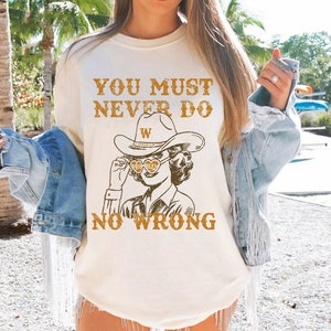 May include: A white t-shirt with the text "You must never do no wrong" and a cartoon image of a woman wearing a cowboy hat and sunglasses. The woman is holding a heart-shaped object with the letter "W" on it.