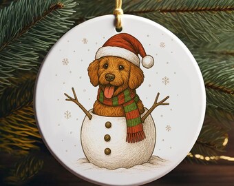 May include: A circular Christmas decoration depicting a golden retriever dog wearing a Santa hat and a green and red scarf, inside a snowman. The ornament is white with snowflakes and a gold hanging string.