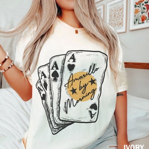 May include: A white t-shirt with a graphic of four playing cards with the text "Amarillo by Morning" in a yellow circle with two stars.