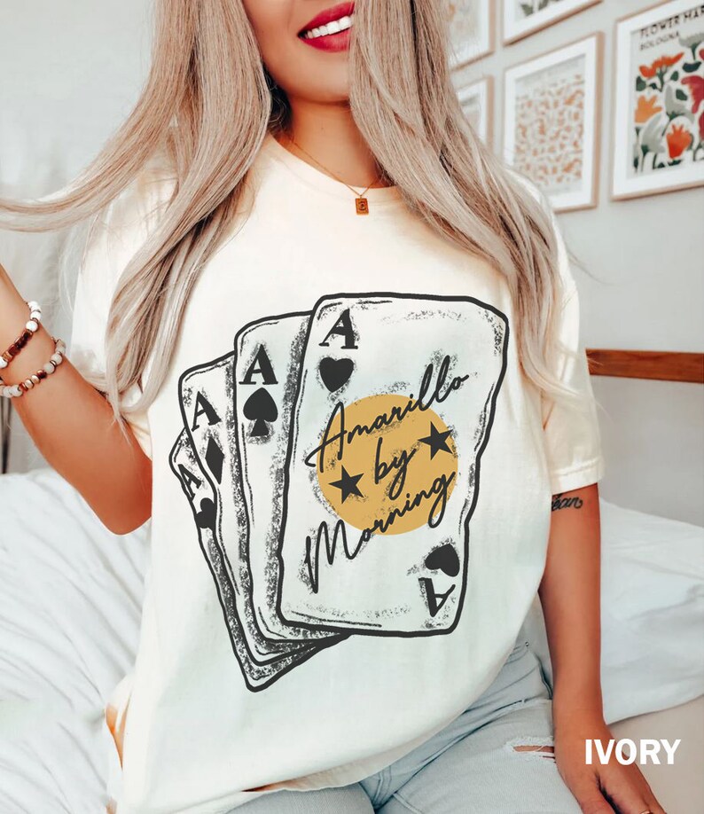 May include: A white t-shirt with a graphic of four playing cards with the text "Amarillo by Morning" in a yellow circle with two stars.