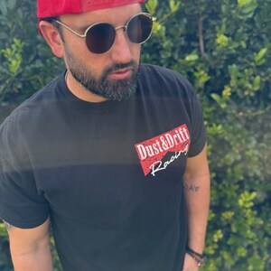 May include: Black t-shirt with the text "Dust & Drift Racing" in red and white. The person in the image is wearing a red trucker hat and round sunglasses.